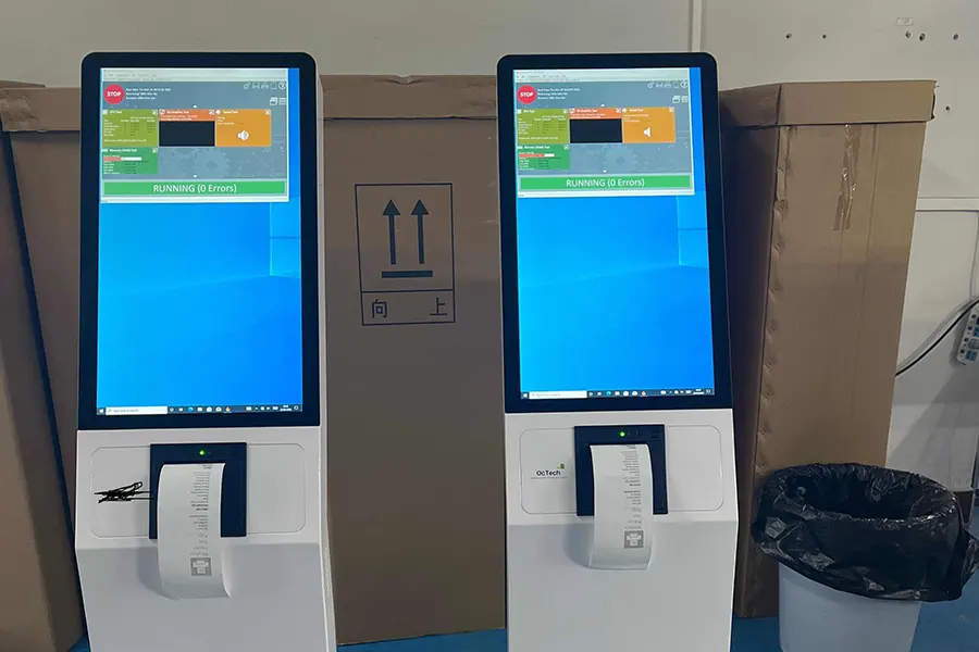 Two floor-standing kiosks powered on with receipts hanging from their printer slots in a warehouse
