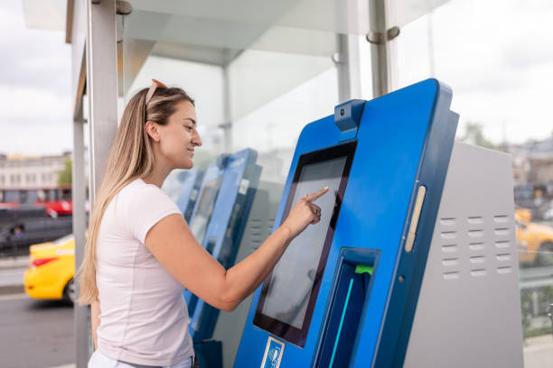 A young woman interacts with a touchscreen on a blue ticket kiosk, demonstrating modern convenience and technology in public transport settings in an urban area.