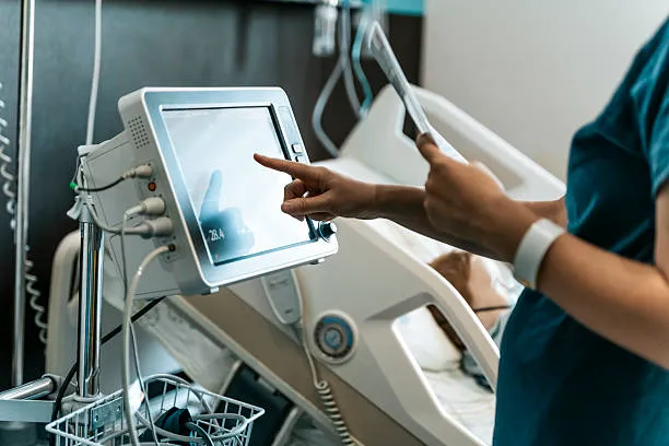 Healthcare Worker's Hand Interacts With A Modern Ventilator Screen In A Hospital Room, Symbolizing Advanced Patient Care And Medical Technology.