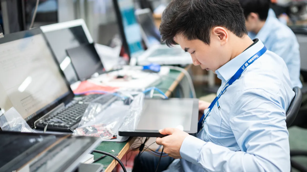 An engineer at a workbench holding a touch panel while a laptop and other displays run behind it