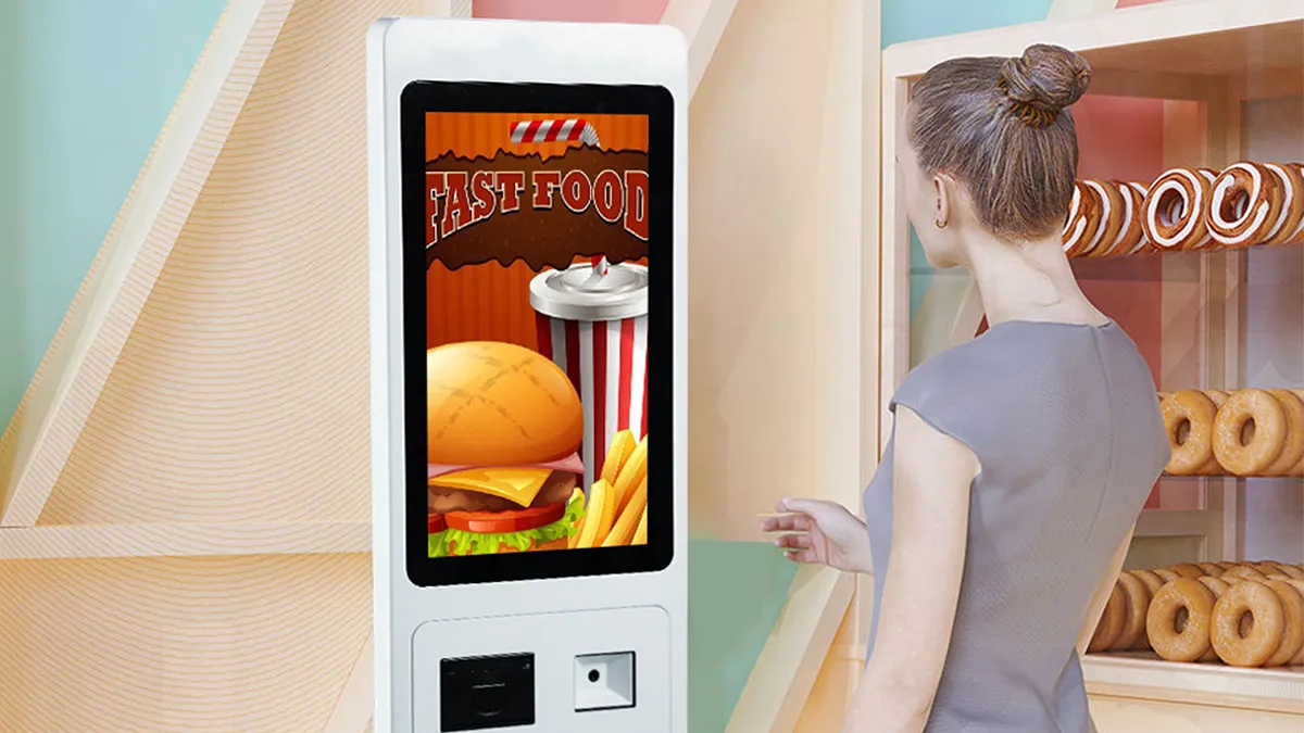 A customer standing beside a white floor-standing ordering kiosk with a fast food menu screen