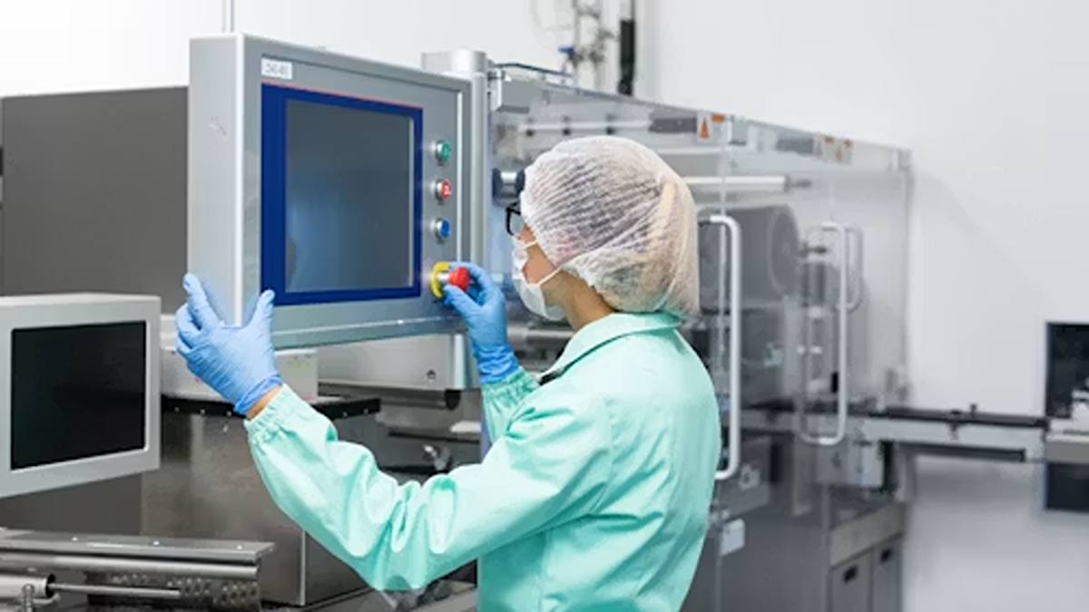 A worker in a cleanroom coat operating a touch control panel mounted on packaging machinery